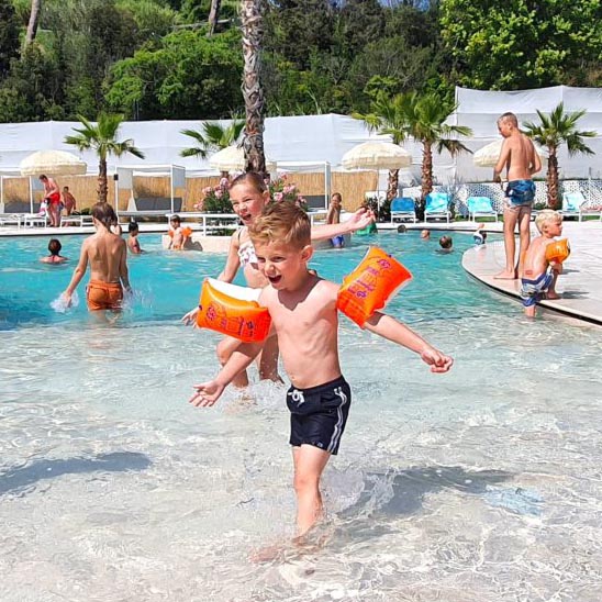 villa-alwin-beach-resorts-kindvriendelijk-zwembad-cupra-marittima Kinderen spelen in het ondiepe zwembad van Villa Alwin Beach Resorts in Cupra Marittima, Italië, met zwembandjes, palmbomen en ligbedden op de achtergrond.