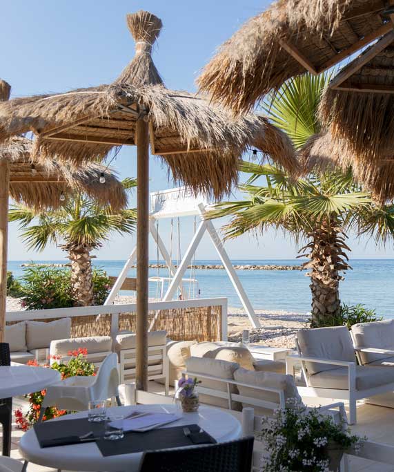 villa-alwin-beach-resorts-beachbar-loungeterras-zeezicht Sfeervol loungeterras van Villa Alwin Beach Resorts in Italië, met comfortabele zitplaatsen, rieten parasols, palmbomen en uitzicht op de Adriatische Zee.